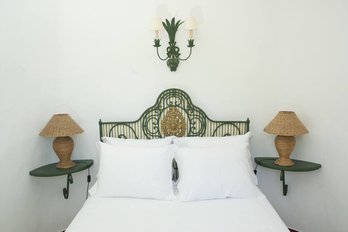 Una cama con cabecera de metal verde, dos lámparas de noche y una lámpara fijada a la pared.