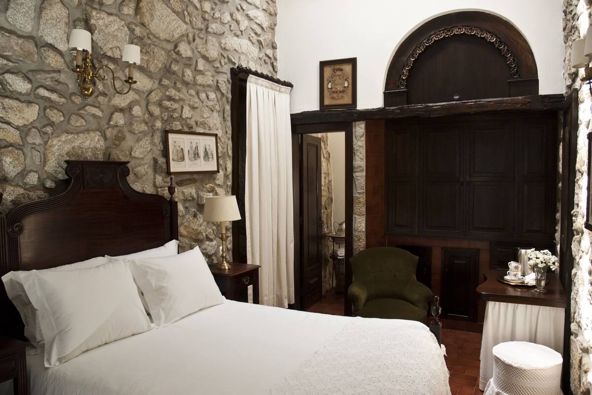 Un dormitorio con pared de piedra, una cama de madera, un sillón verde y una mesita con un jarrón de flores.
