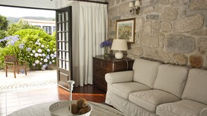 A living room with a stone wall, a sofa, a table with fruit, and a view of a garden with flowers. - Casa do Ameal (Viana do Castelo)
