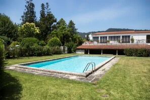 A rectangular swimming pool with a stone border, surrounded by a well-maintained lawn and various trees. - Casa do Ameal (Viana do Castelo)