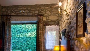 A bedroom with a stone wall, a bed, a nightstand with a lamp, and a view of greenery outside. - Casa do Ameal (Viana do Castelo)