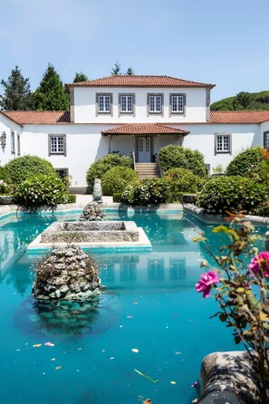 A white building with a tiled roof, a swimming pool with a fountain, and surrounding greenery.