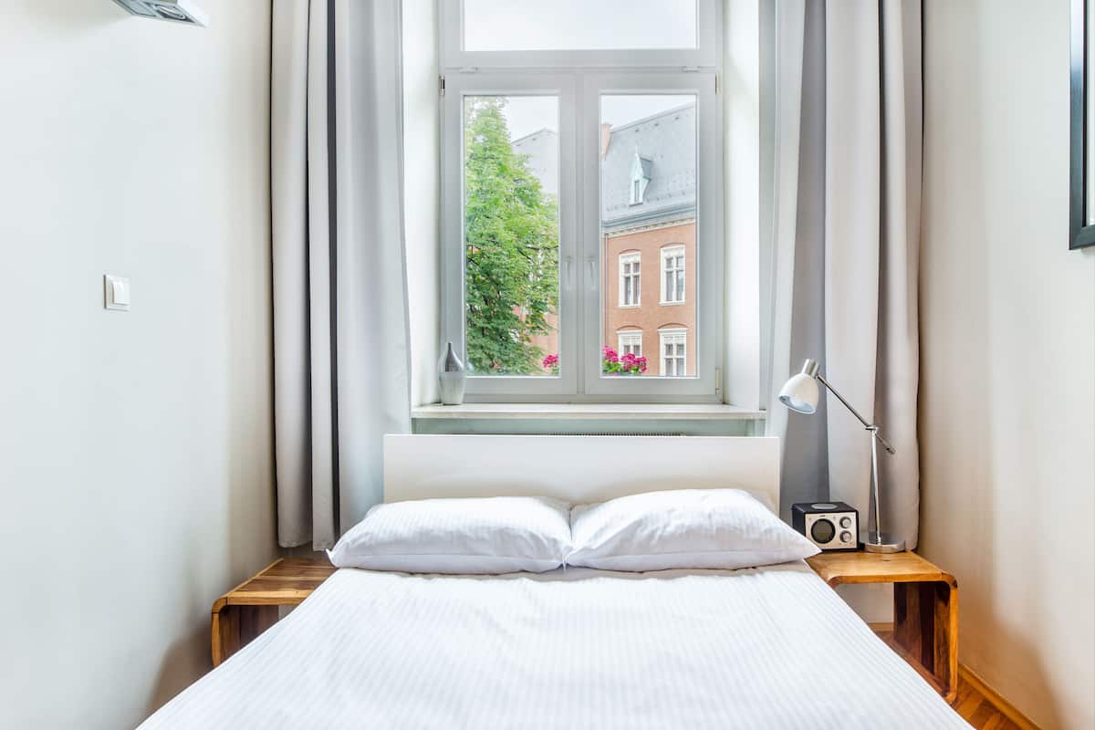 Una cama bien hecha con sábanas blancas, una mesita de noche con lámpara y radio, y una ventana con cortinas.