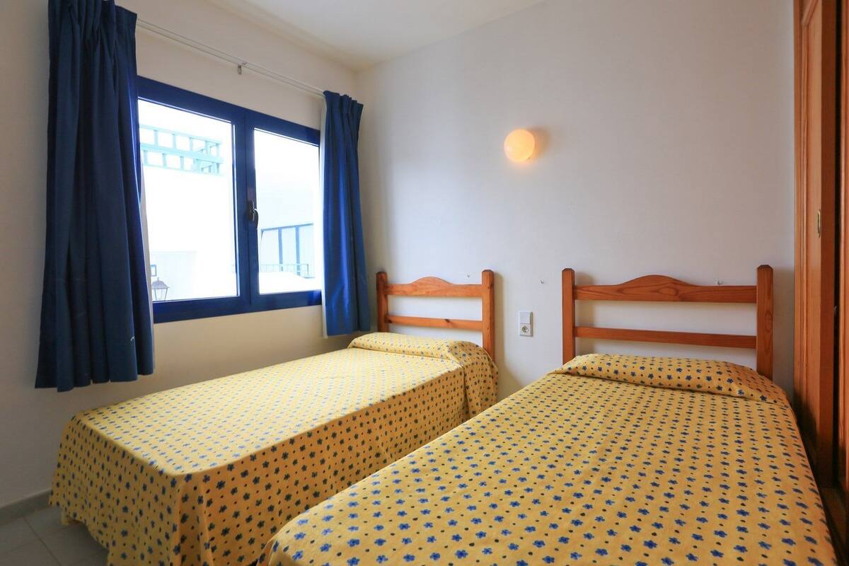 A room with two single beds, a window with curtains, and a wall-mounted light.