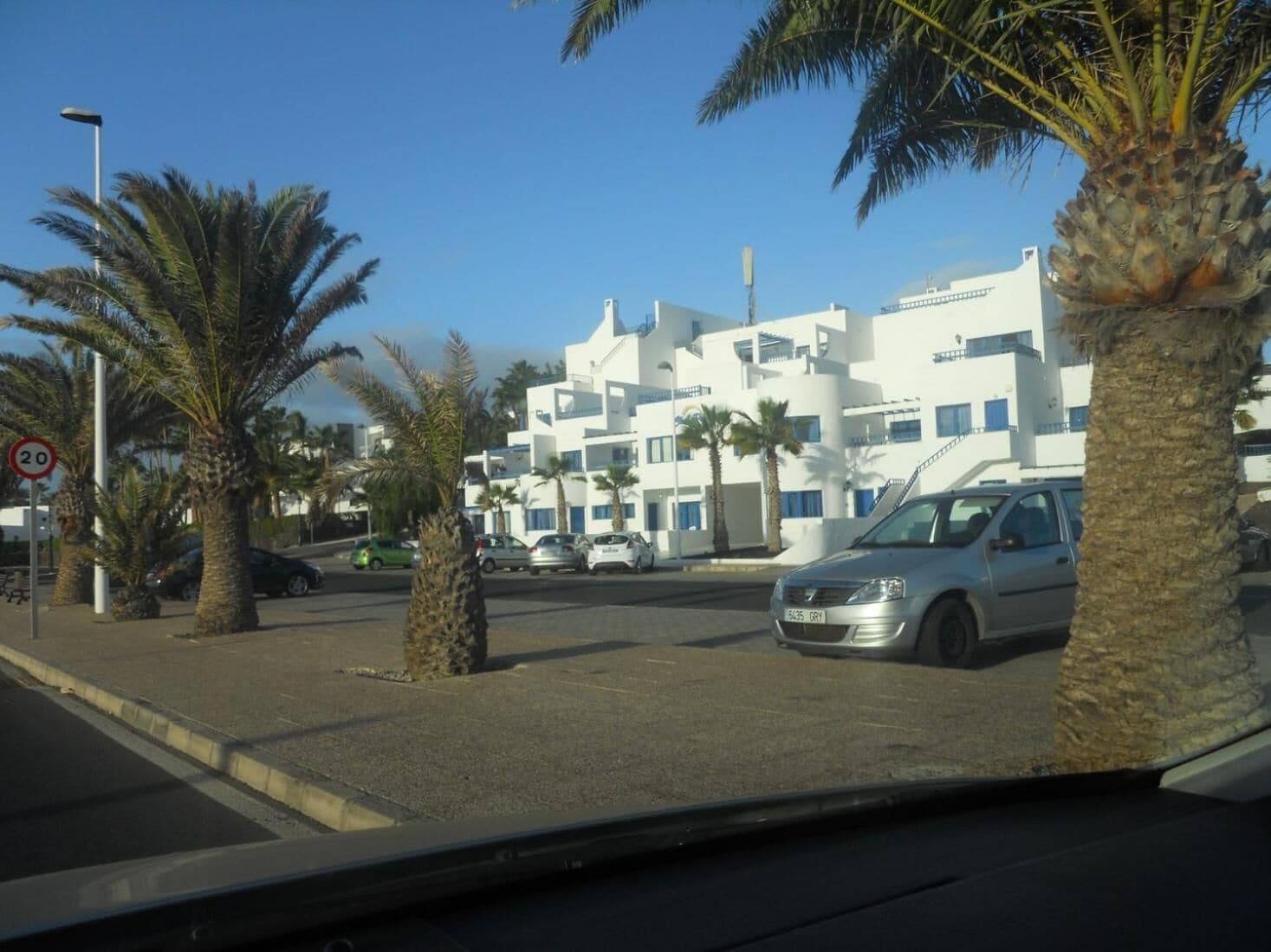 A modern building with white exterior and balconies, surrounded by palm trees and parked cars.