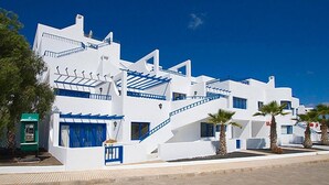 A white building with blue accents, including stairs and railings, and palm trees in front. - Apartamentos Club Pocillos (Tías)