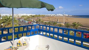 A balcony with a green umbrella, white chairs, and a table with a menu. A beach and cityscape are visible in the background. - Apartamentos Club Pocillos (Tías)
