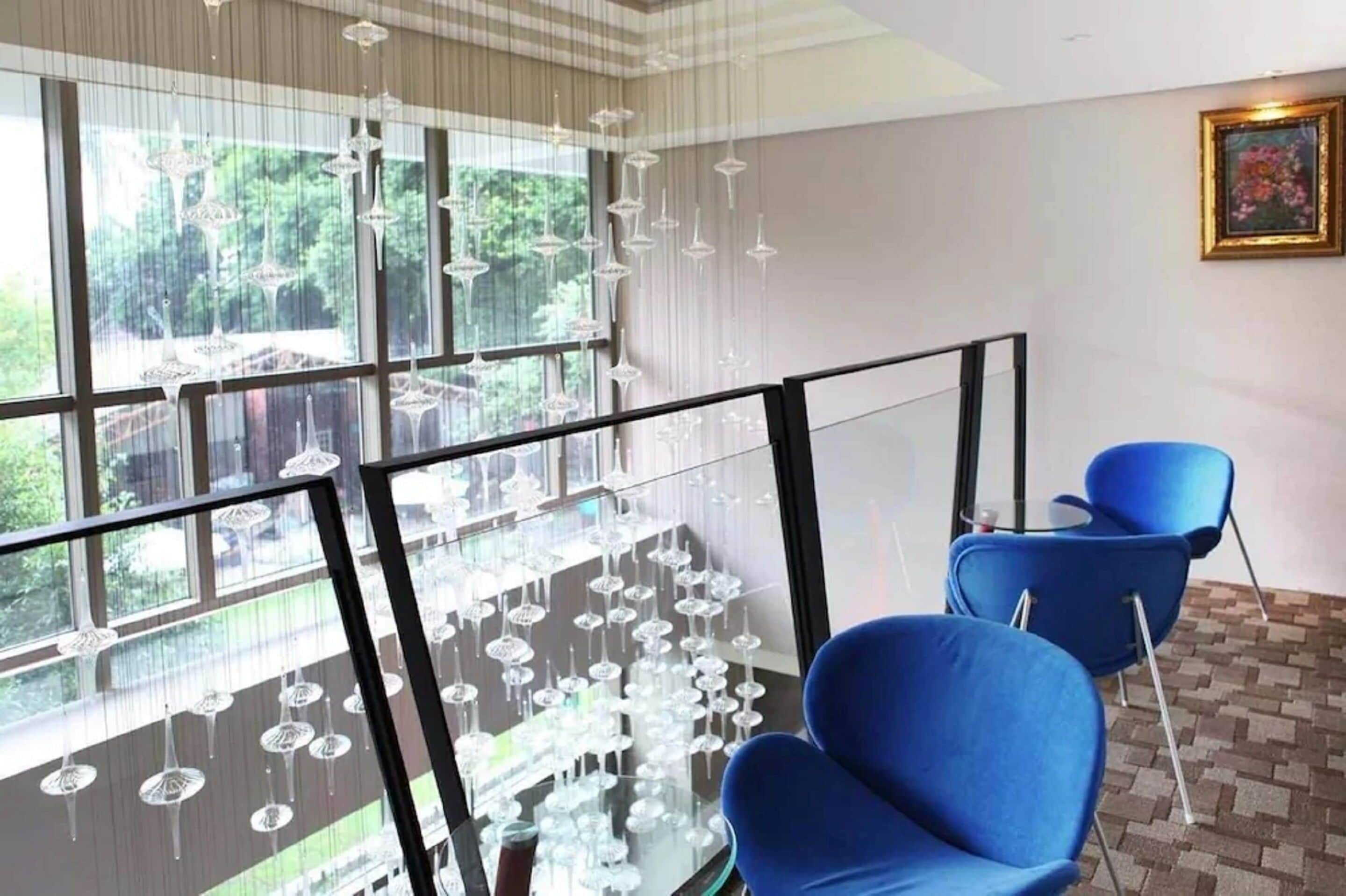 A modern hotel room with large windows, a balcony with hanging lights, and blue chairs.