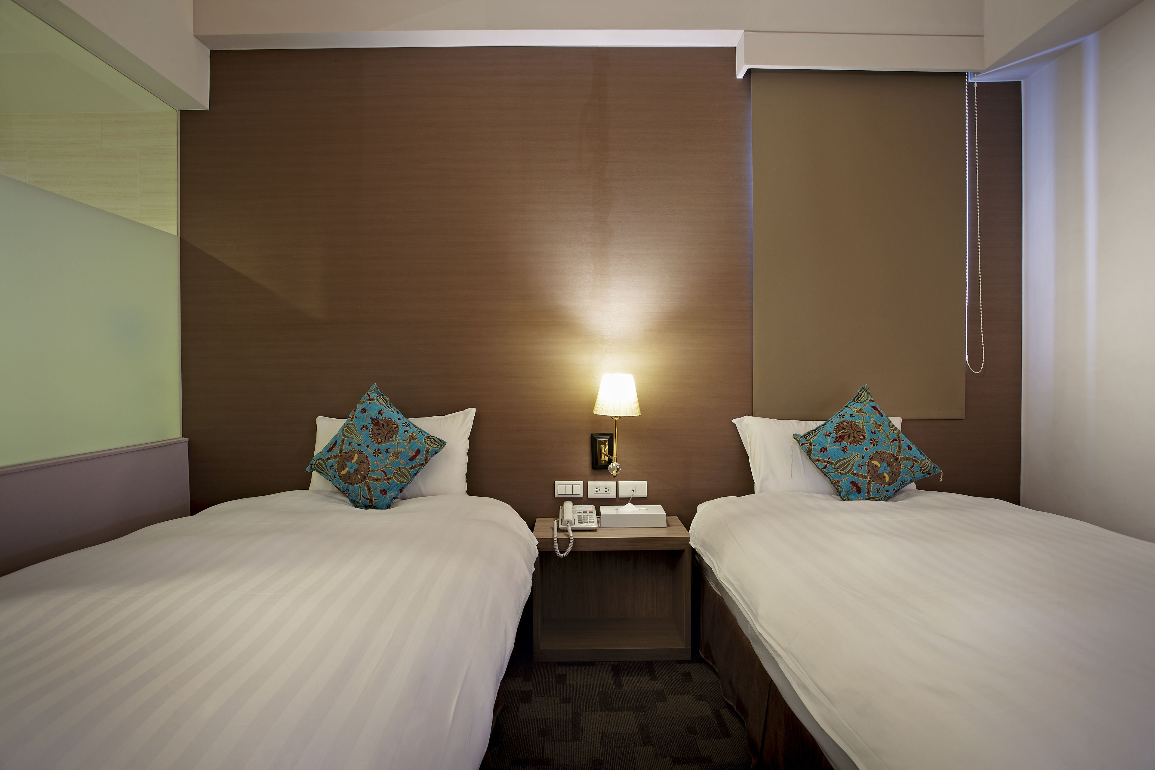 Habitación de hotel con dos camas, una mesita de noche con un teléfono y una lámpara, y un cabecero de madera.