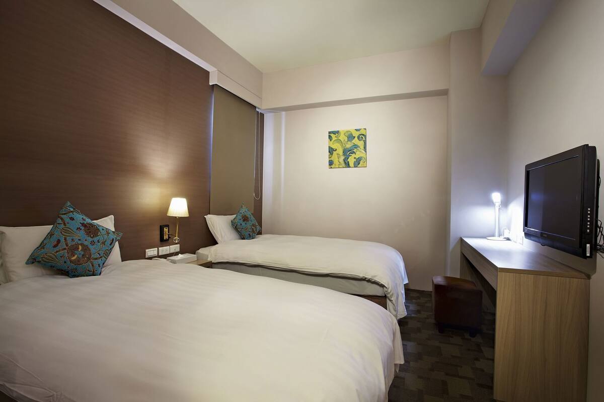 A hotel room with two beds, a desk with a TV, a lamp, and a painting on the wall.