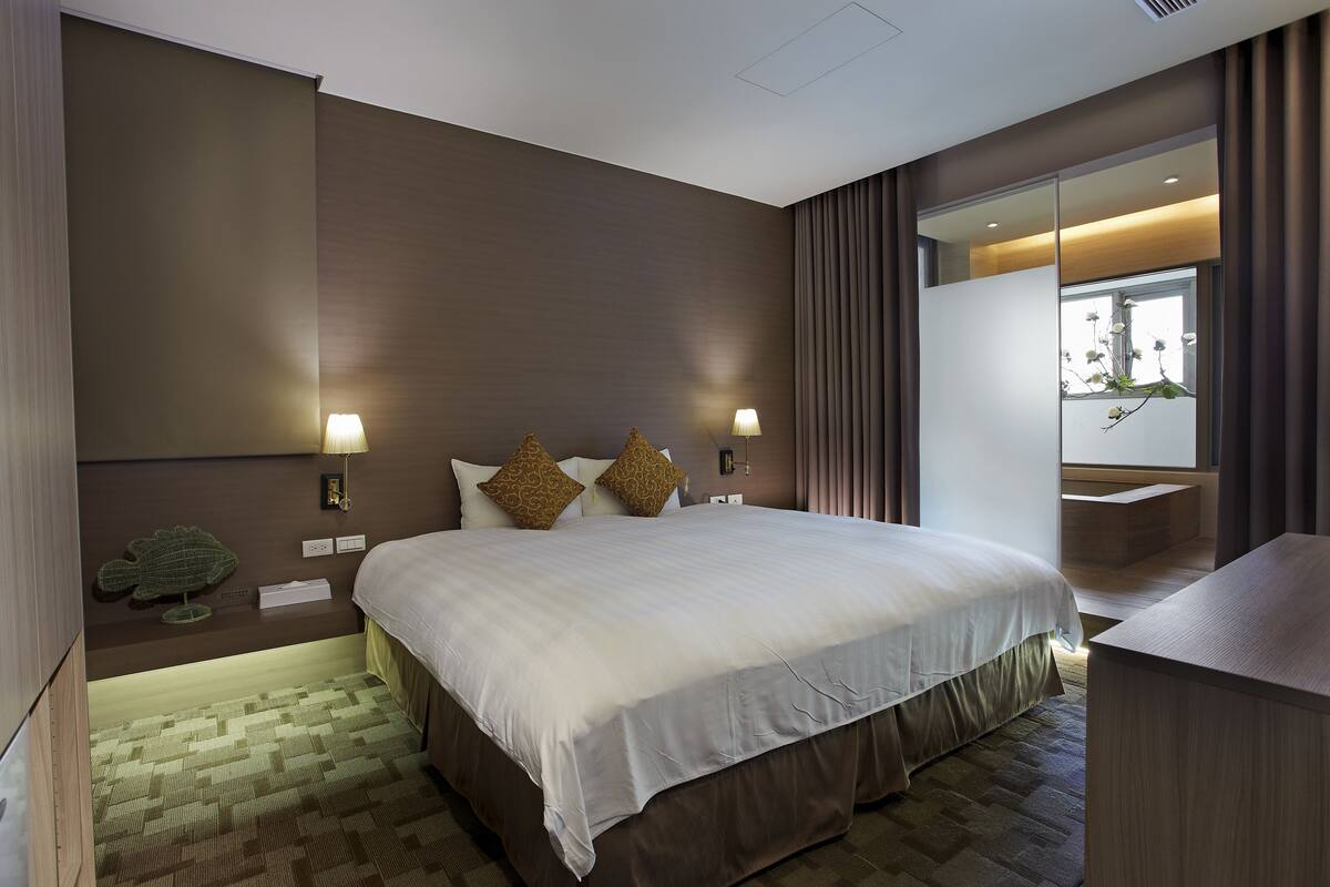 A modern hotel room with a large bed, two bedside lamps, a small side table, and a view of the outdoors through a sliding door.