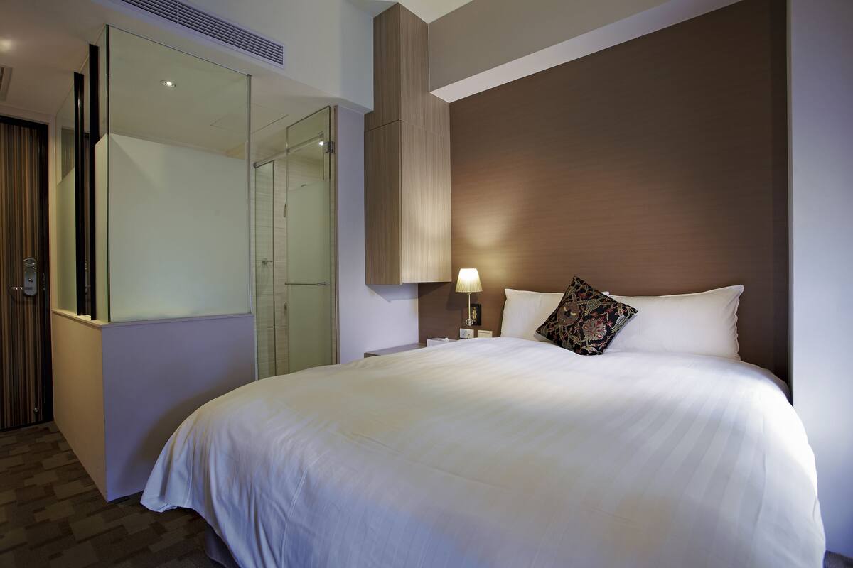 A hotel room with a large bed, a nightstand with a lamp, a glass shower enclosure, and a wardrobe.