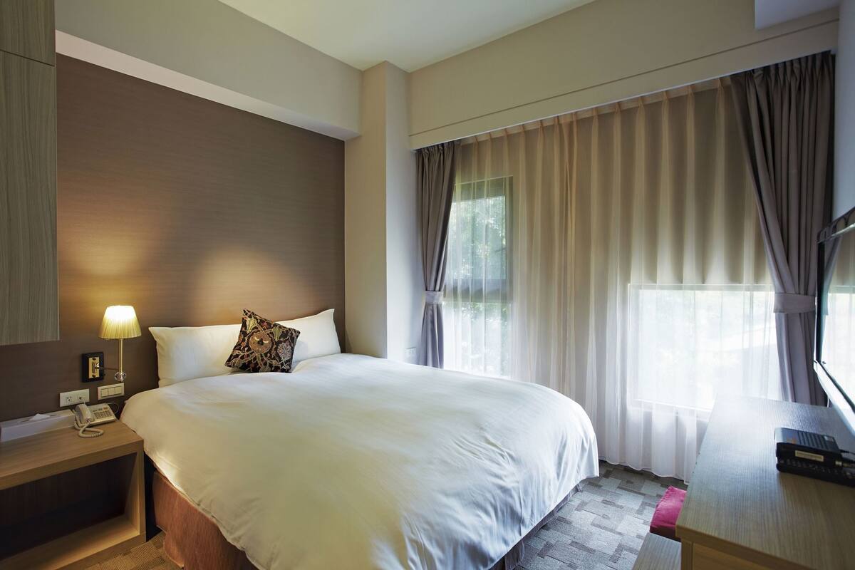 A hotel room with a large bed, bedside table with a lamp, a telephone, and a TV on a wooden table.