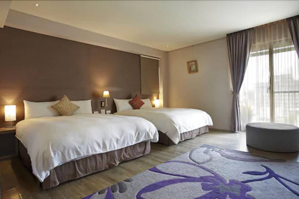A hotel room with a large bed, a bedside table, a lamp, a window with curtains, and a small ottoman.