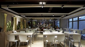 Un comedor moderno con mesas y sillas blancas, ventanas amplias y un techo con vigas y luminarias a la vista.
