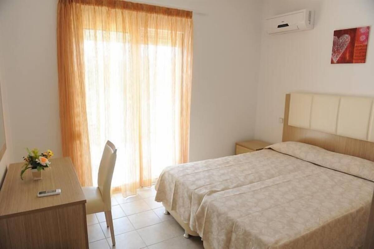 A hotel room with two beds, a desk with a chair, a window with curtains, an air conditioner, and a wall-mounted artwork.