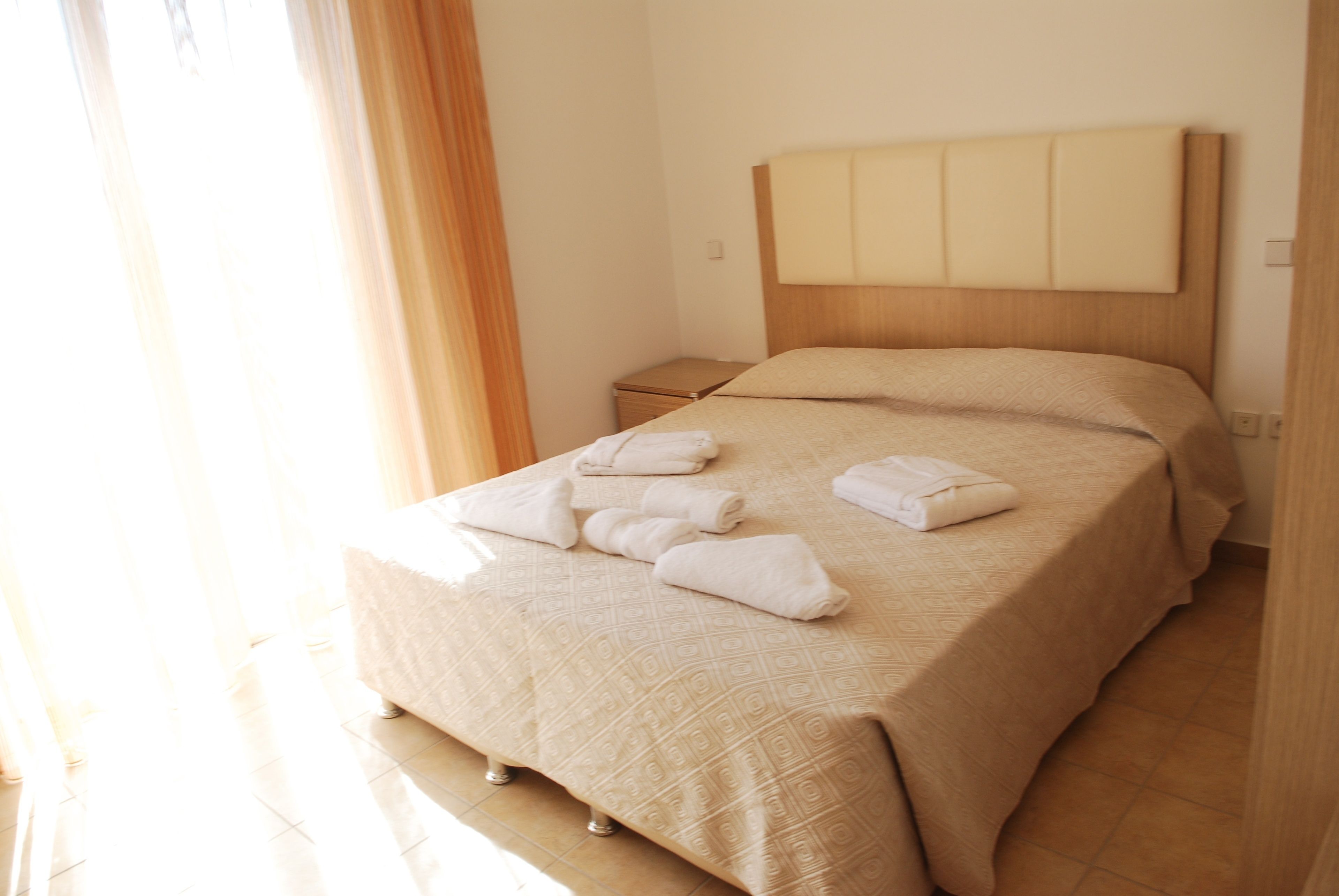 A neatly made bed with a headboard, a bedside table, and a window with curtains.
