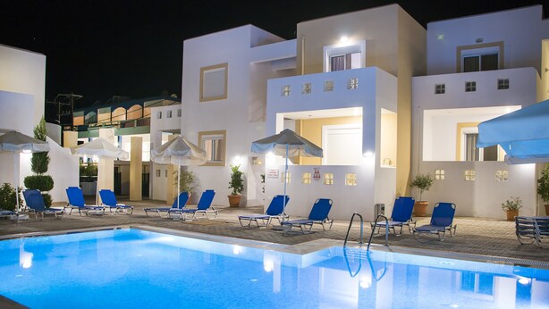 Una piscina con agua azul, rodeada de edificios blancos con numerosas ventanas y puertas. Hay sillas de descanso azules y sombrillas alrededor de la piscina.