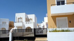 Una hilera de edificios modernos, en tonos blanco y beige, con balcones, un acceso con portones y un cielo azul despejado.