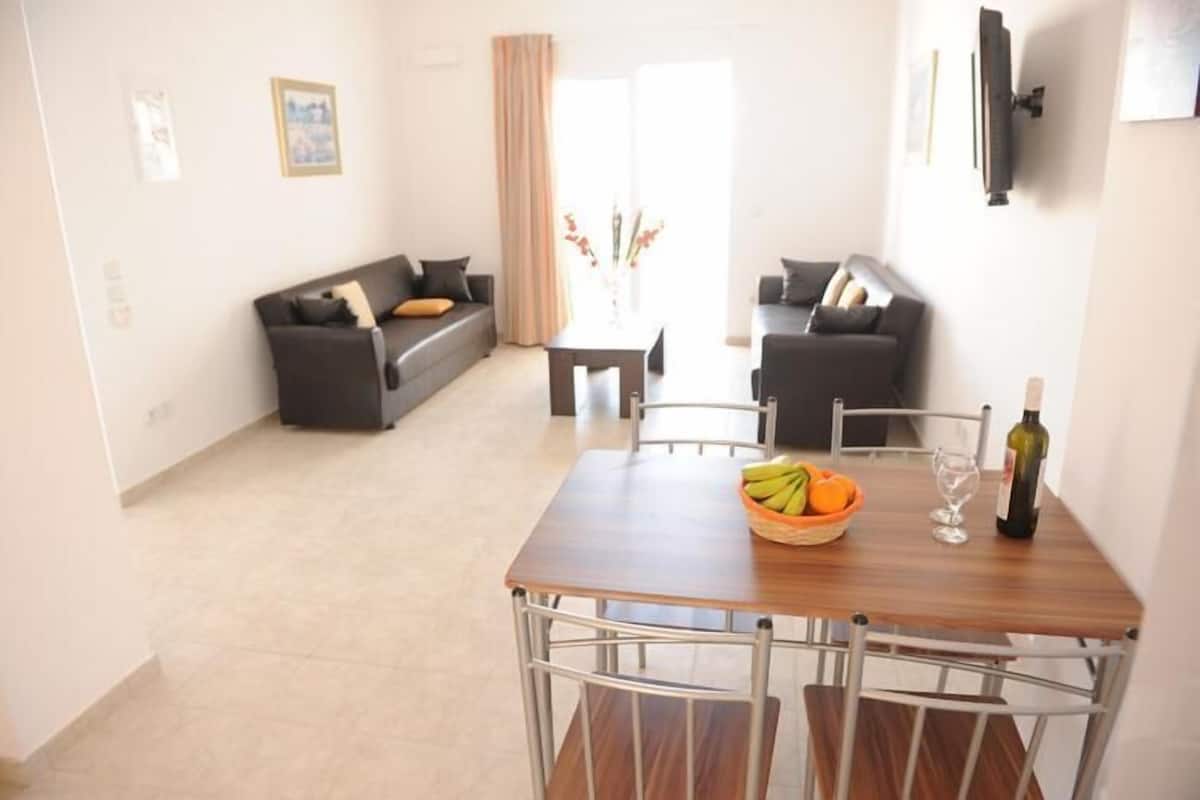 A modern living room with a sofa, a dining table with chairs, and a TV mounted on the wall.