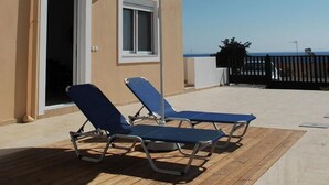 Un edificio con fachada color crema, una terraza de madera, dos sillones azules y un paraguas color crema.