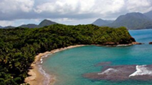 Un paisaje costero con una playa, aguas turquesas cristalinas y colinas verdes exuberantes al fondo.