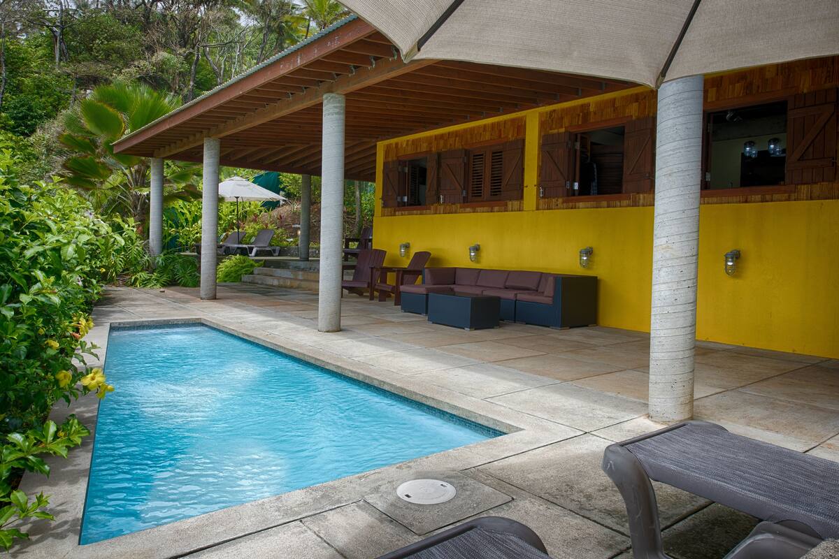Un espace aménagé au bord de la piscine, avec un coin salon couvert, une piscine et un mur jaune.