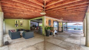 Un área de estar al aire libre con techo, que ofrece vistas al océano, con vigas de madera, piso de piedra y una zona de bar.
