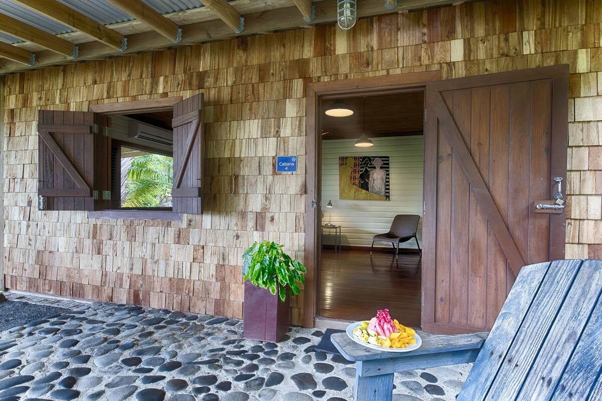 Un bâtiment rustique en bois, doté d’une entrée pavée de pierres, d’une plante en pot et d’une table où sont disposés des aliments.