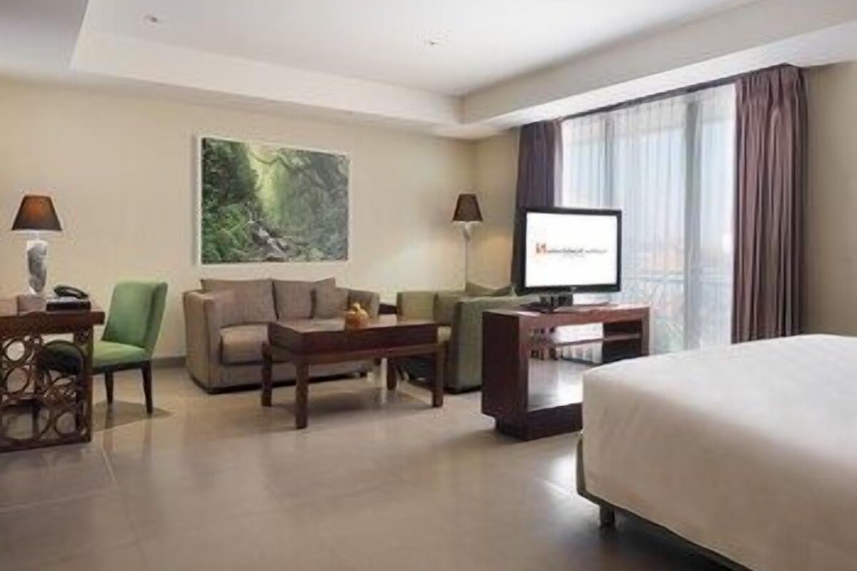 A hotel room with a bed, a desk with a computer, a chair, a sofa, a TV, and a painting on the wall.