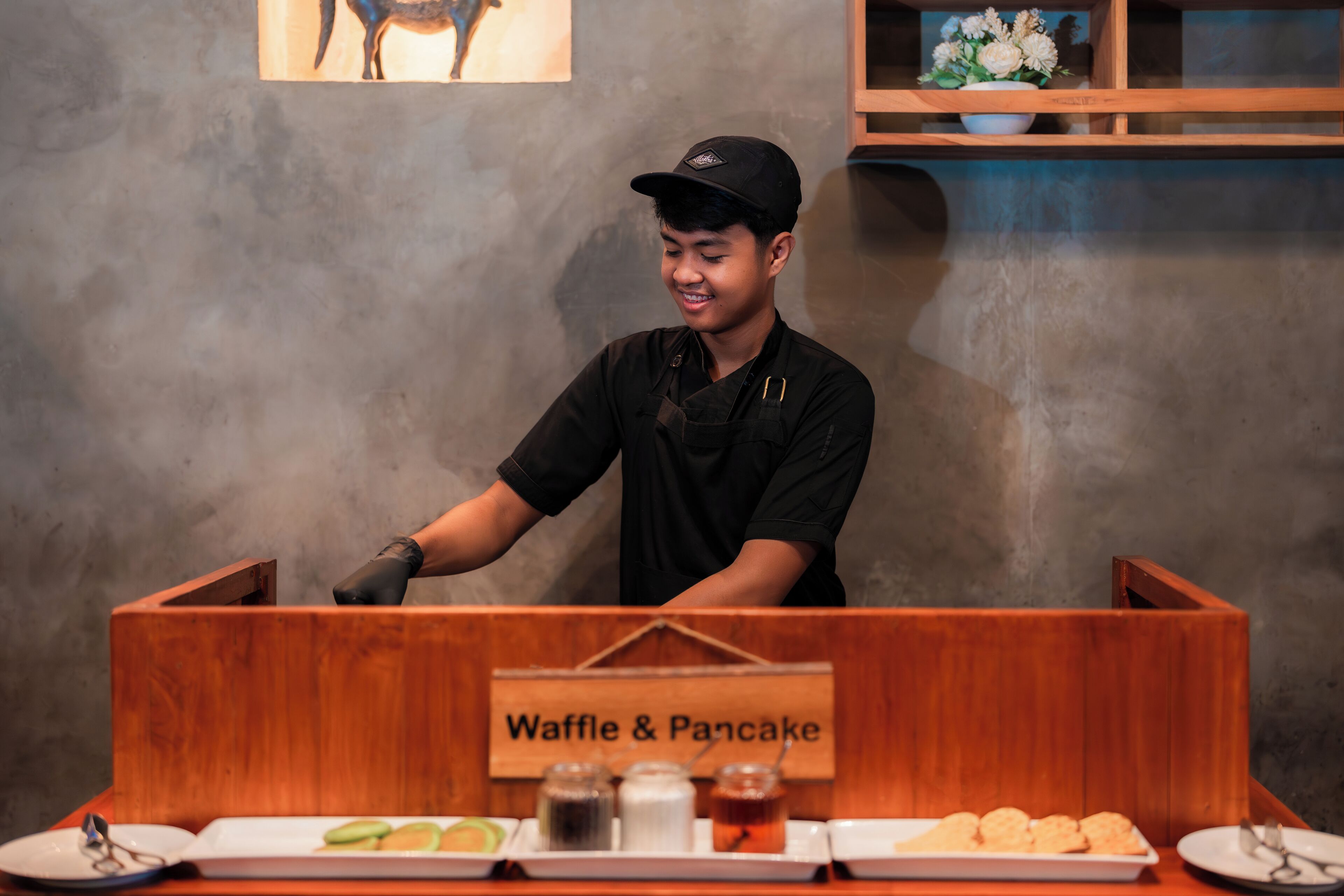 A person serving food behind a counter labeled "Waffle & Pancake" with various dishes and condiments.