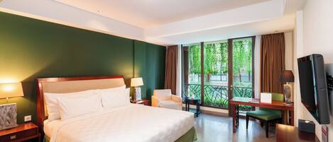 A hotel room with a large bed, a desk with a chair, a television, and a view of greenery outside.
