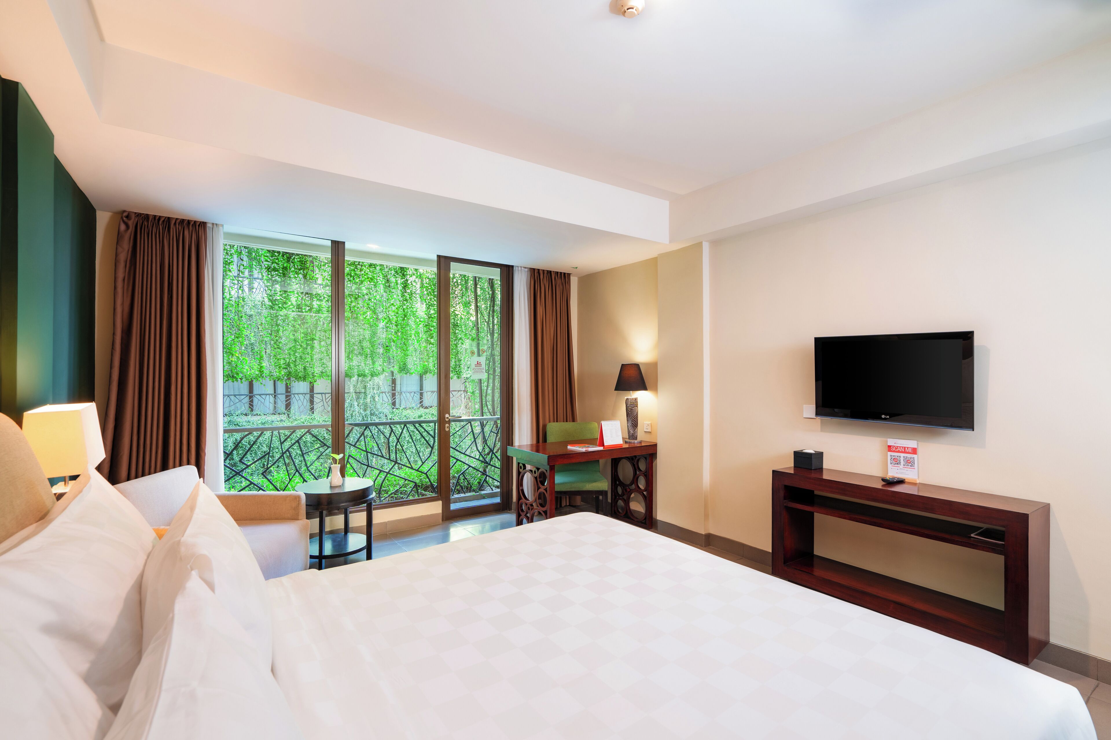 A hotel room with a large bed, a desk with a chair, a television, and a balcony with a view of greenery.