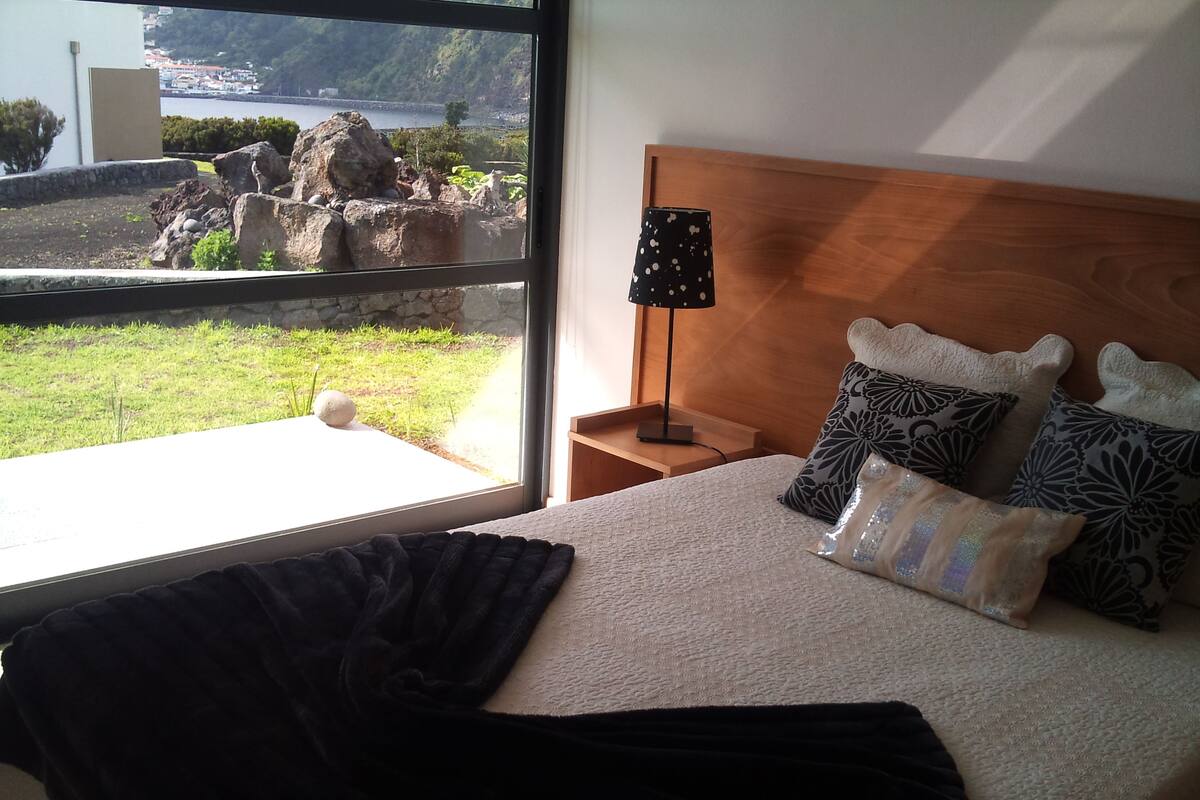 Un dormitorio con un ventanal grande, una cama con almohadas estampadas, una mesita de noche y una lámpara.
