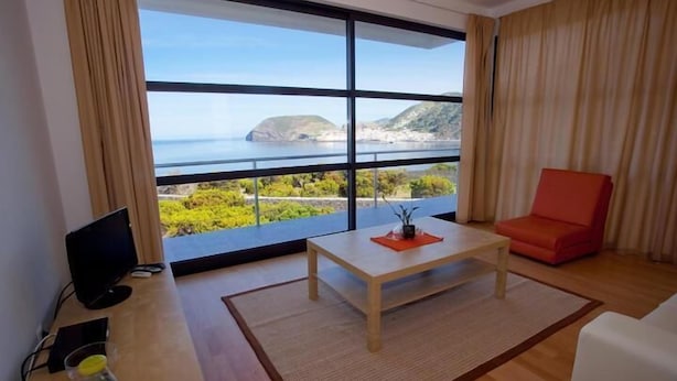 Un salon moderne doté d’une grande fenêtre offrant une vue panoramique sur la mer et les collines.