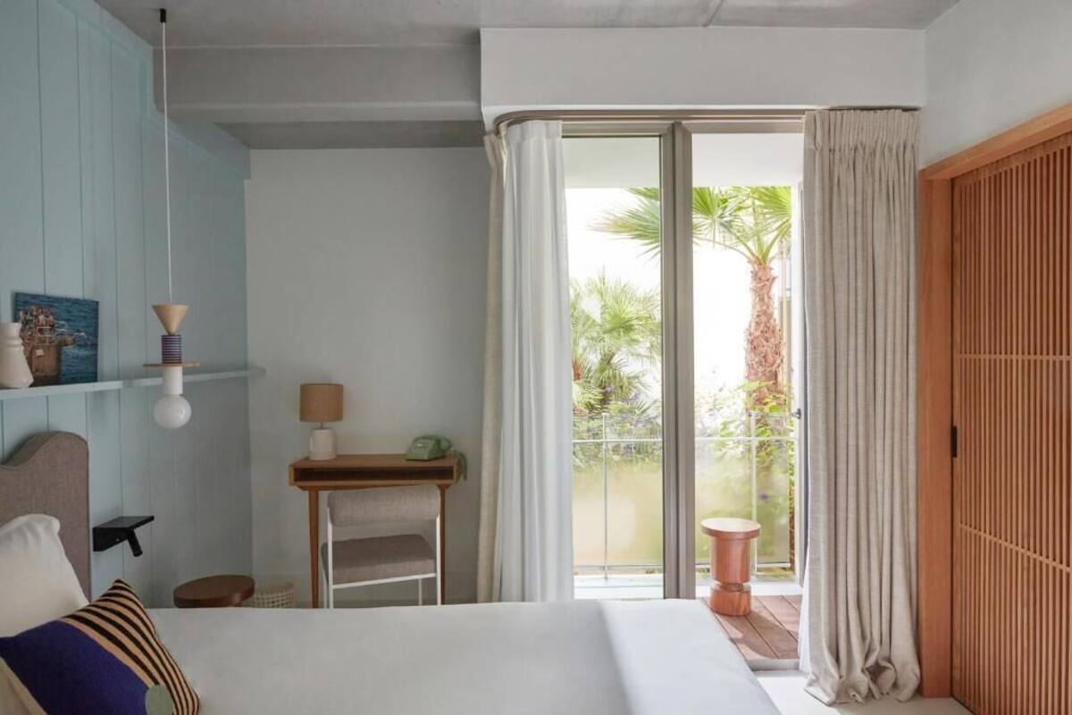 Ein Hotelzimmer mit Bett, kleinem Tisch mit Lampe, Stuhl, Holzschrank und einer Glastür, die auf einen Balkon mit Pflanzen führt.