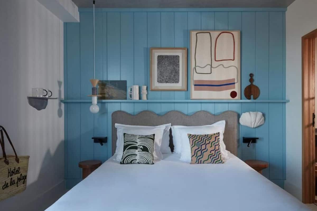 Ein Schlafzimmer mit blauer Wand, einem Bett mit zwei Kissen, einem gerahmten Bild und einer kleinen Ablagefläche mit Gegenständen.