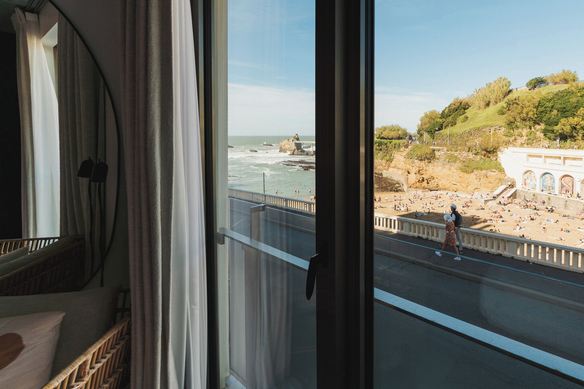 A view from a window overlooking a beach with people, a road, and a building with arches.