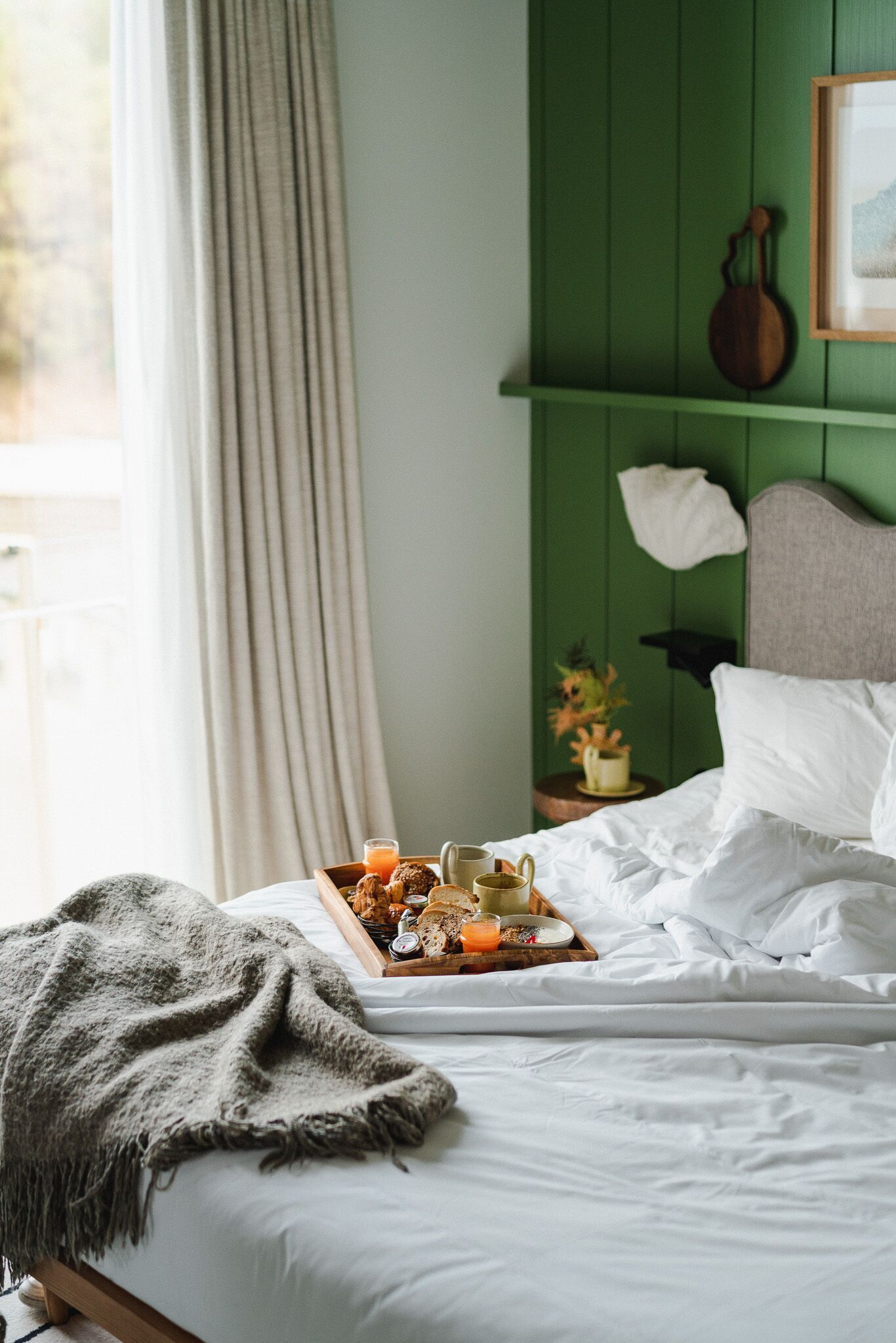 A neatly made bed with a tray of breakfast items, a green wall, and a window with curtains.