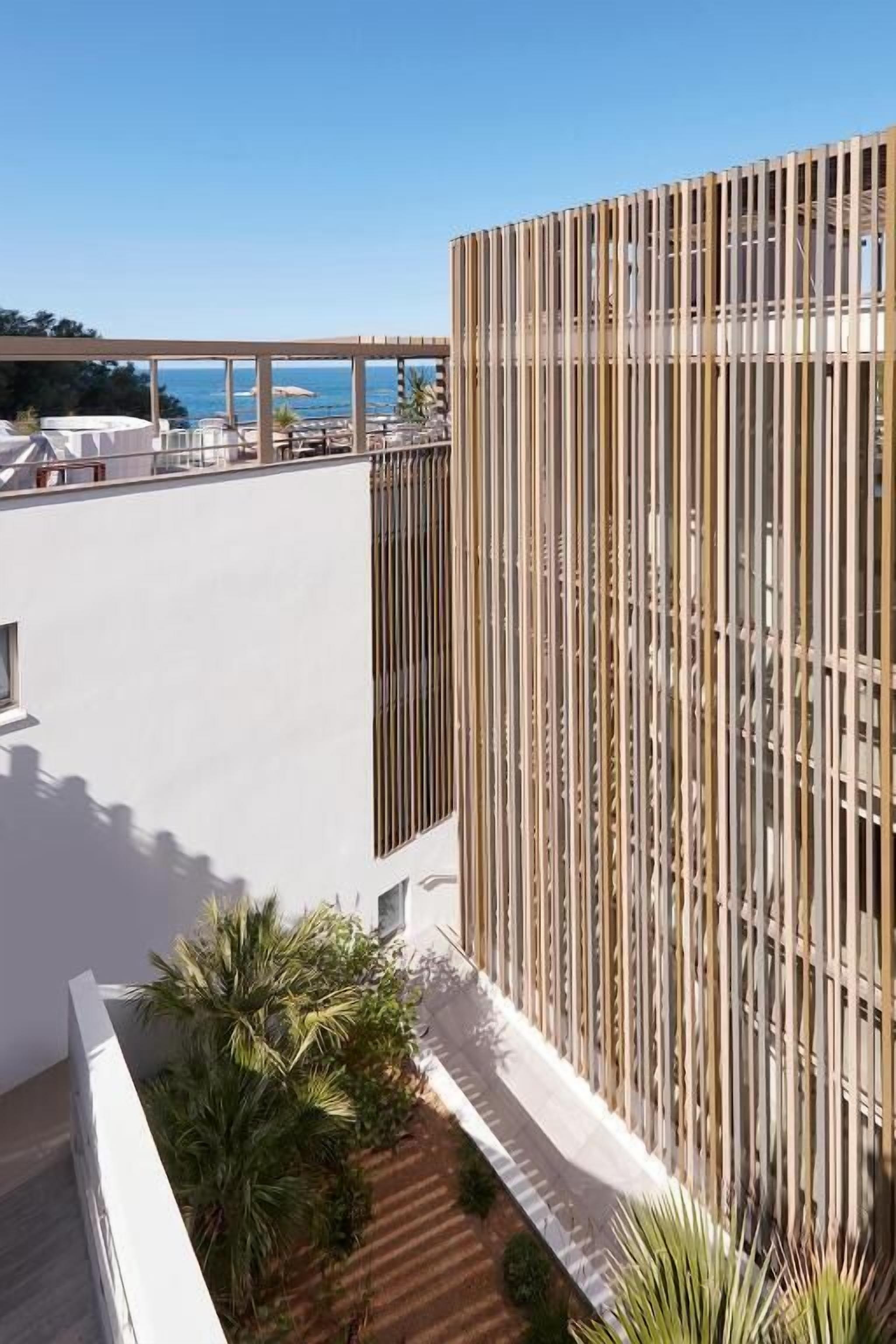 Un bâtiment moderne doté d’une façade verticale en bois et d’une terrasse sur le toit, aménagée avec des chaises longues et des parasols.