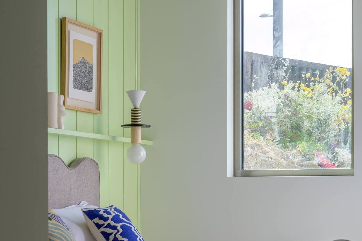 Ein Schlafzimmer mit einem Bett, einem Sessel, einem Fenster mit Blick ins Grüne und einer Wandleuchte.
