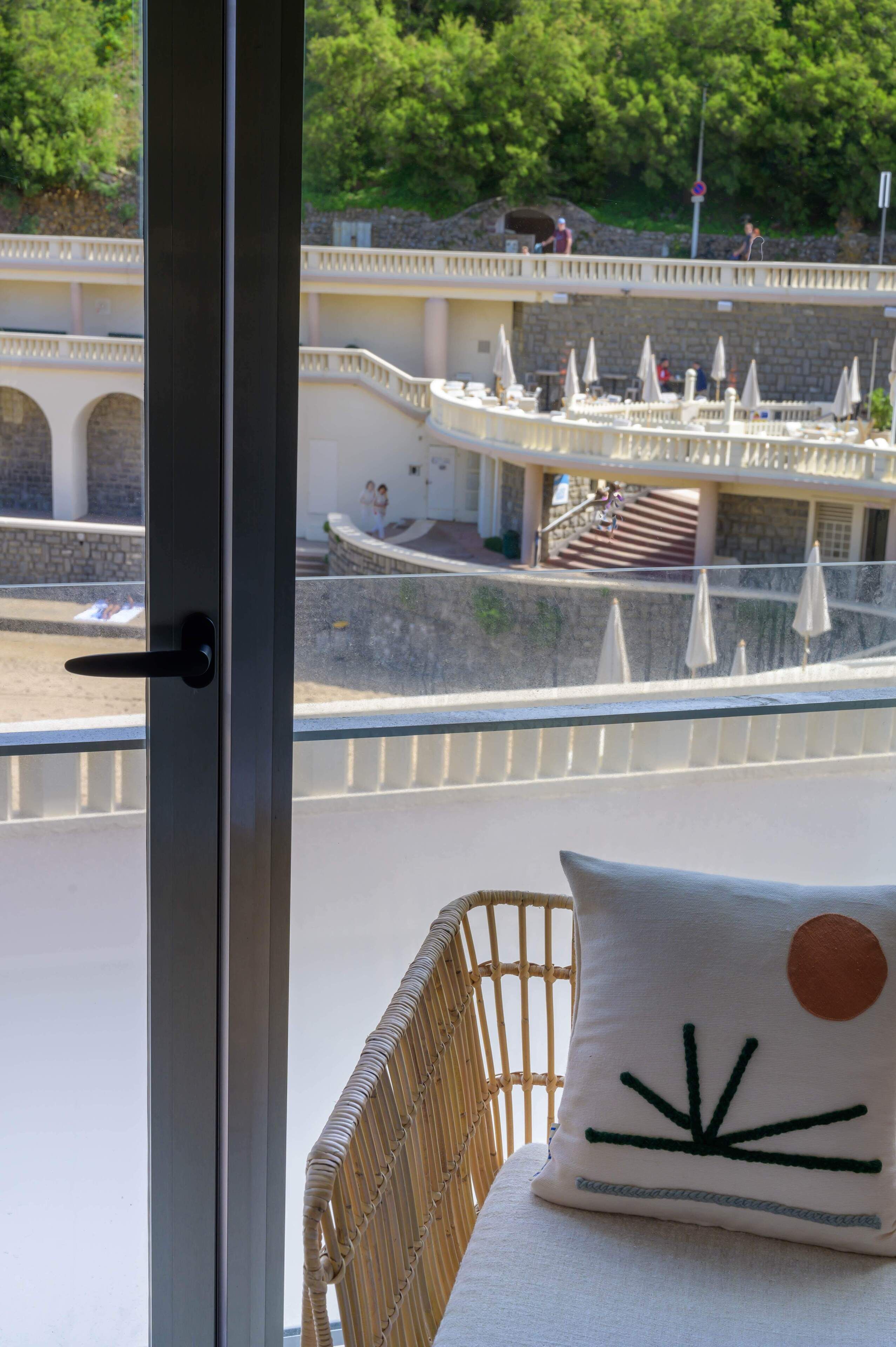 Un balcón con una silla de mimbre y un cojín con diseño de sol, con vistas a una zona de piscina con sombrillas y un muro de piedra.