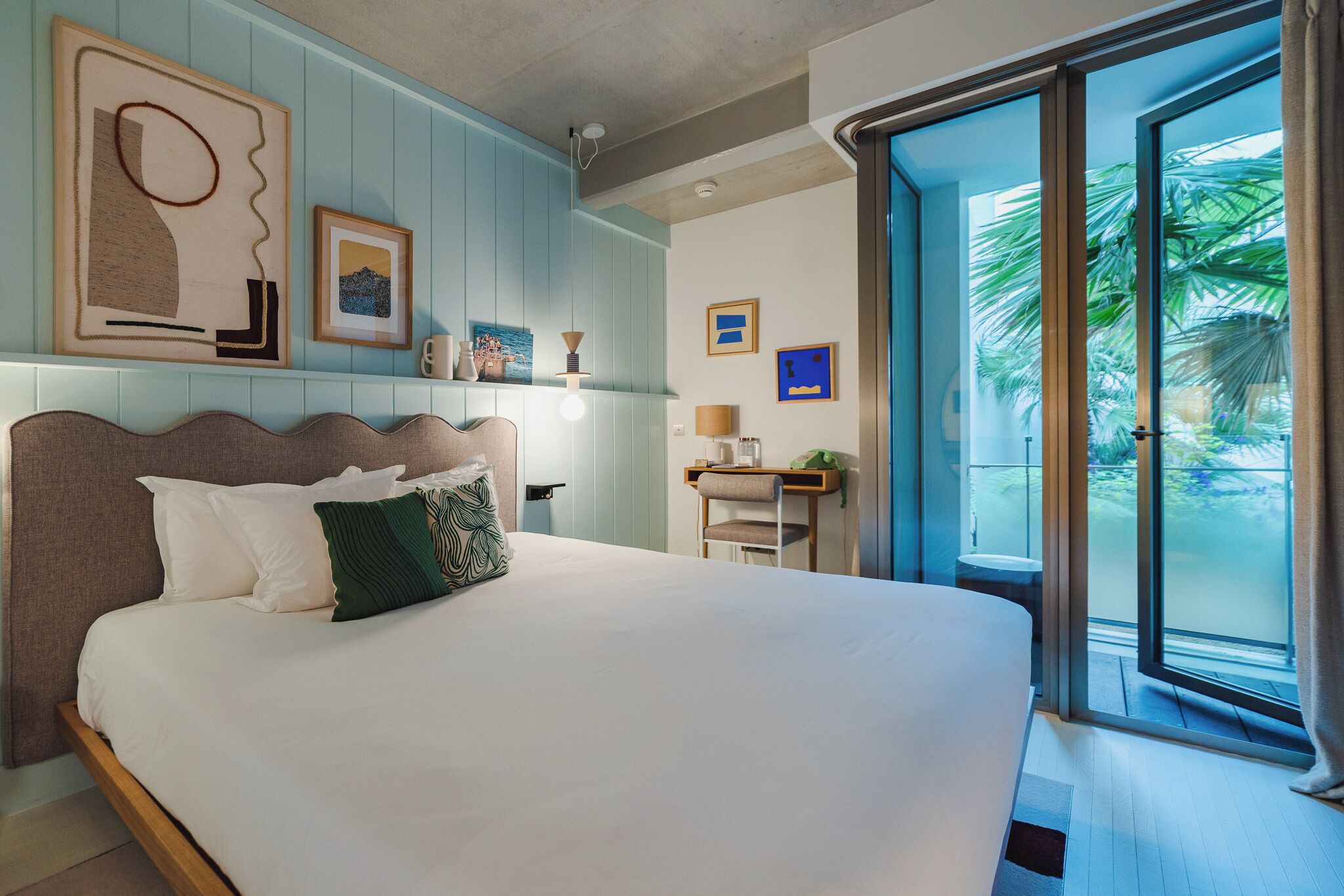 A hotel room with a large bed, a desk, and a view of palm trees through a sliding glass door.