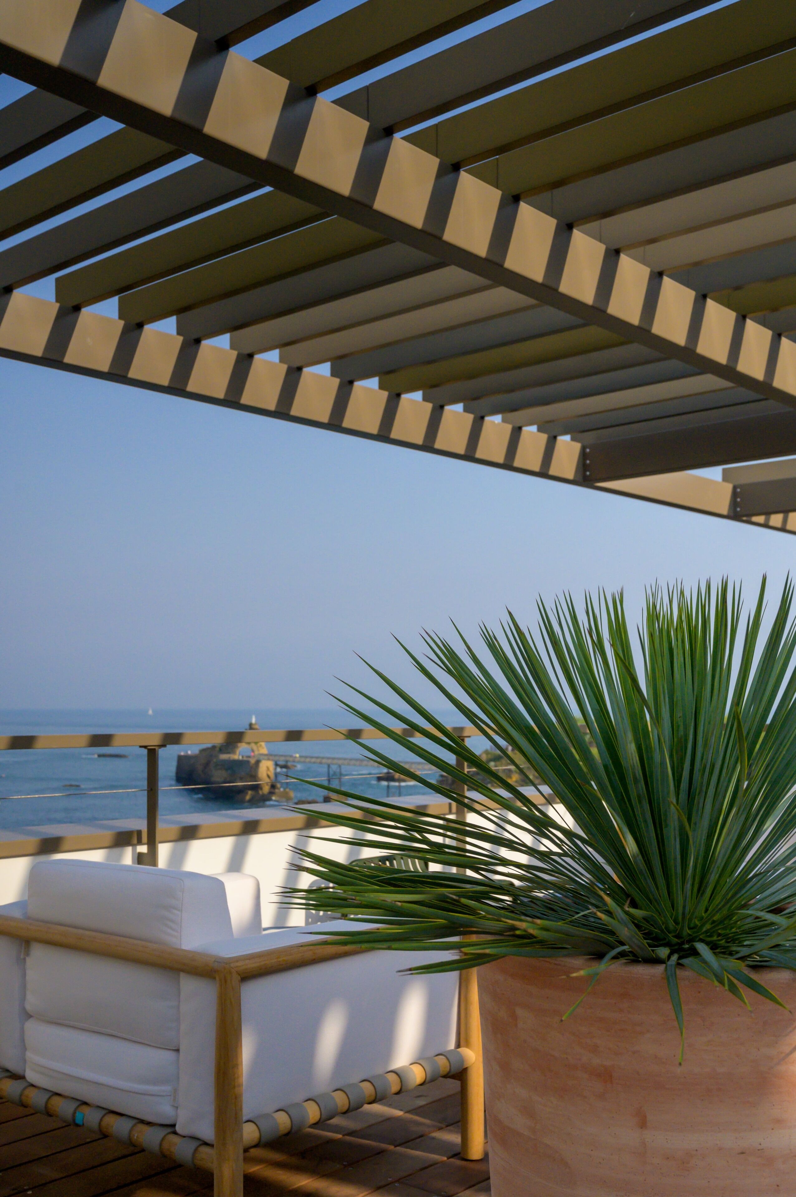 A covered outdoor seating area with a view of the ocean, a potted plant, and a white cushioned chair.