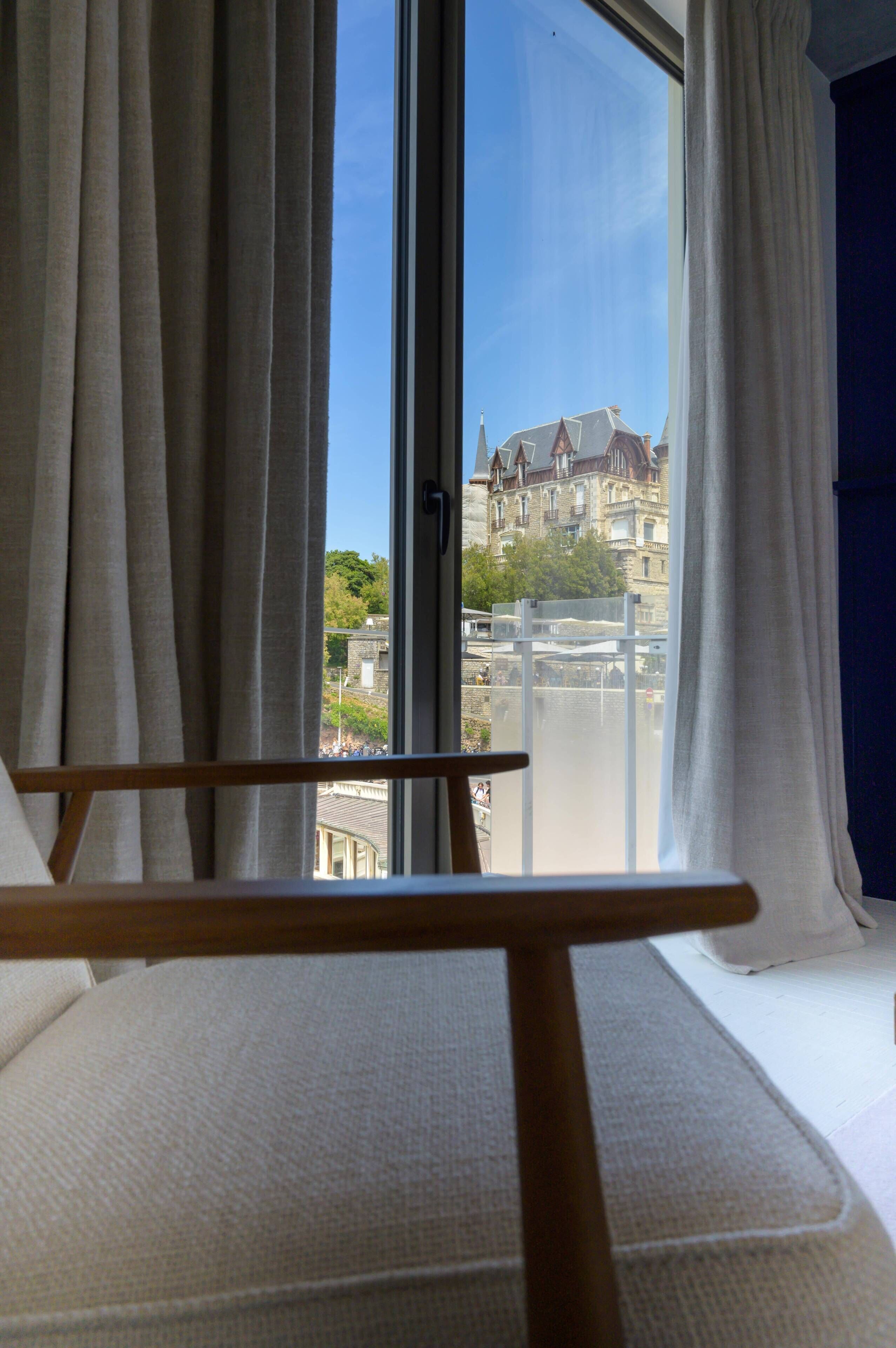 A room with a view of a building through a window with curtains.