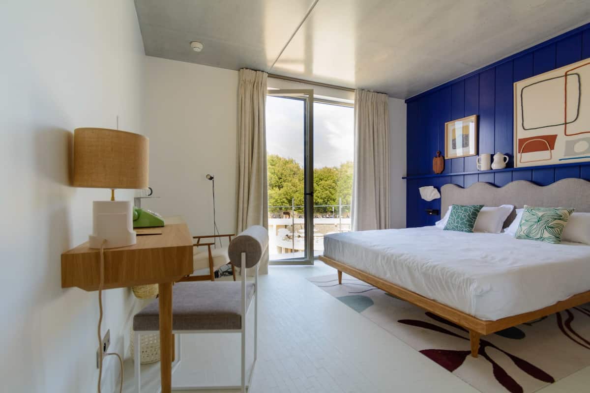 Ein modernes Schlafzimmer mit Bett, Nachttisch, Lampe, Sessel und Blick ins Freie.