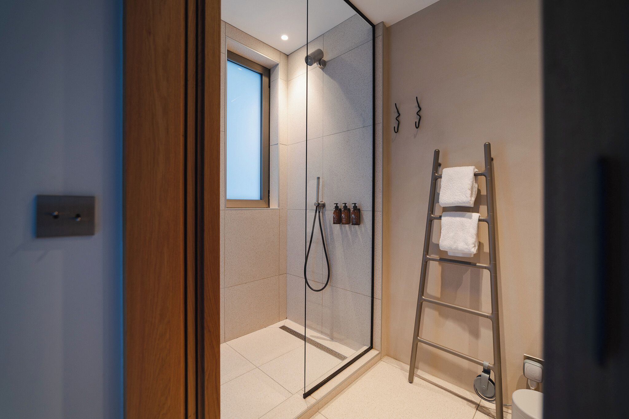 A modern bathroom with a glass shower enclosure, a wooden door, a small window, and a towel rack.