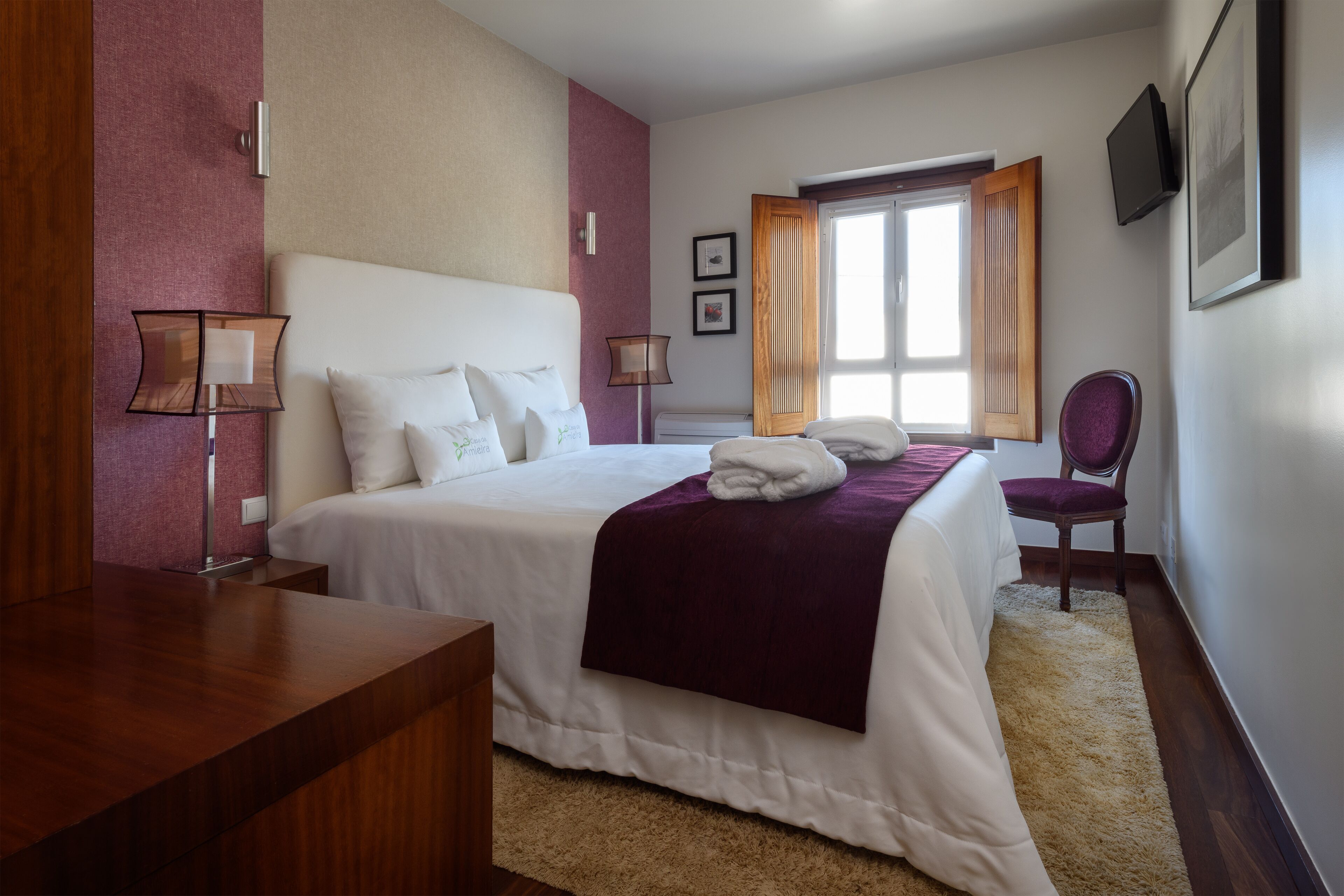 A hotel room with a large bed, a wooden desk, two bedside lamps, a chair, and a window with curtains.