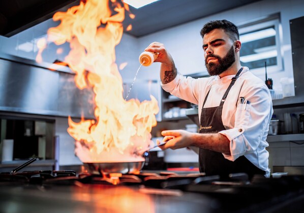Ein Koch bereitet Essen auf einem Herd zu, umgeben von Flammen in einer professionellen Küche.