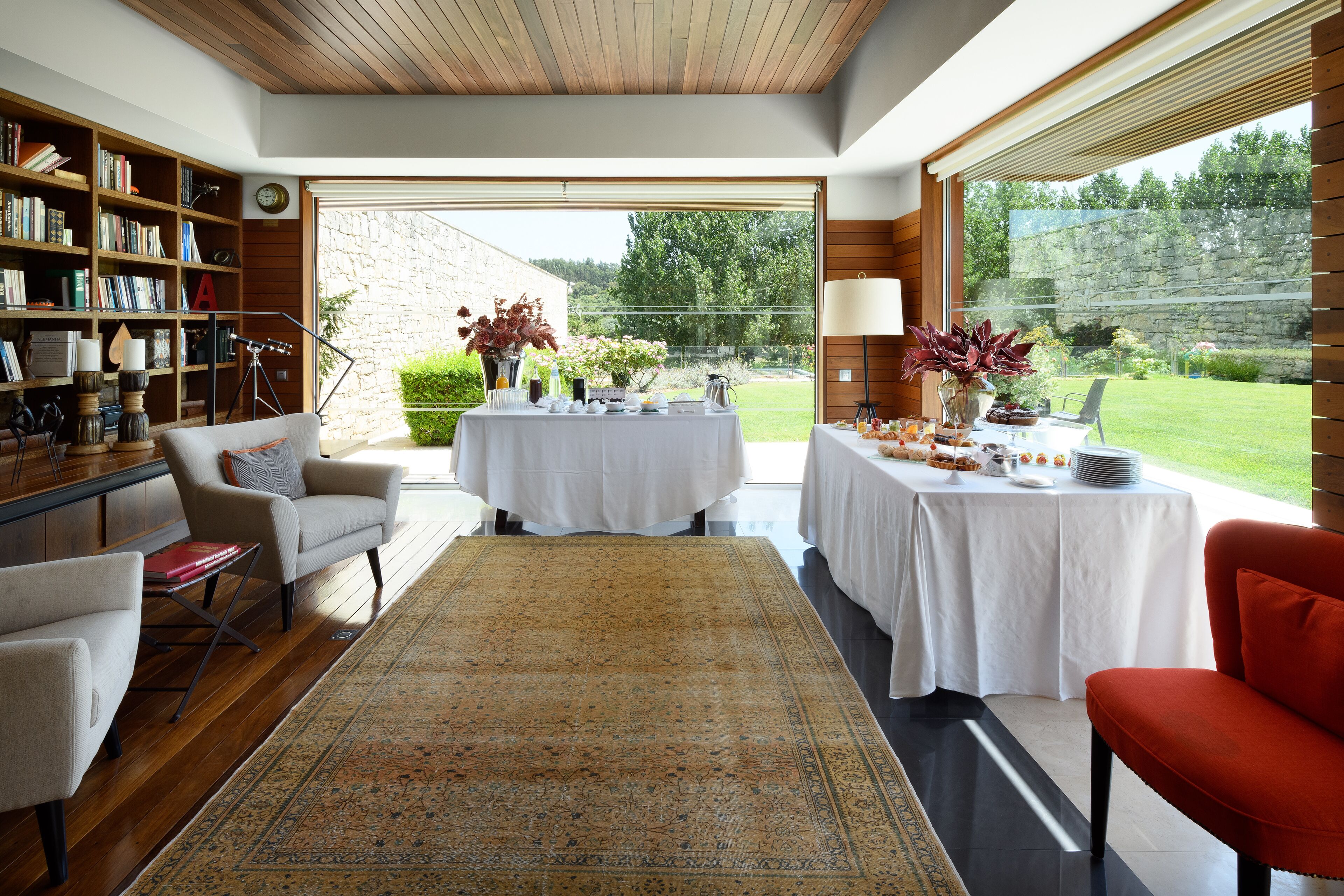 A spacious living area with a large window, a bookshelf, a dining table set for a meal, and a red chair.